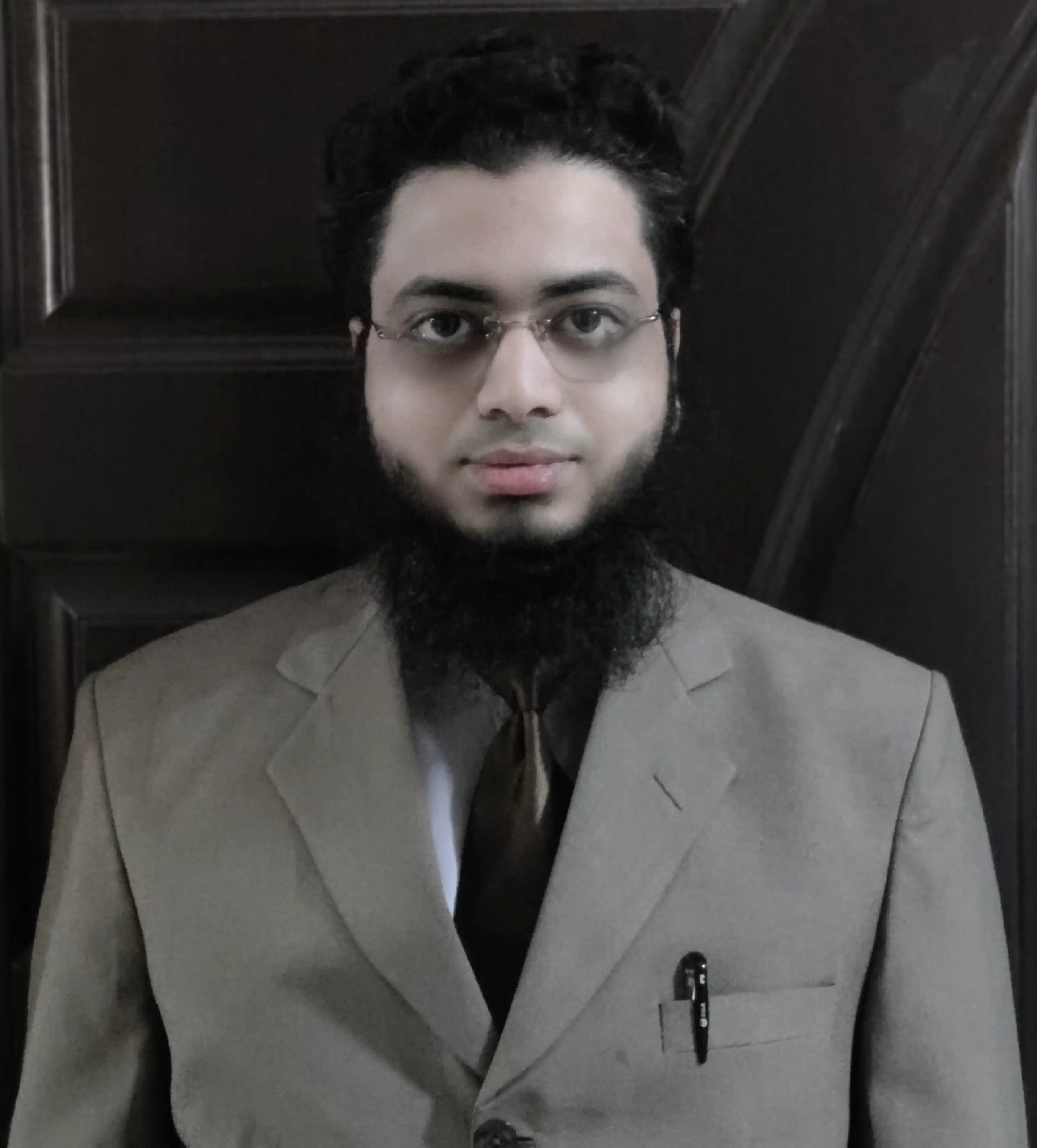 Professional photo of Wajeeh Misbah Khan in a beige suit and tie against a dark wooden background.